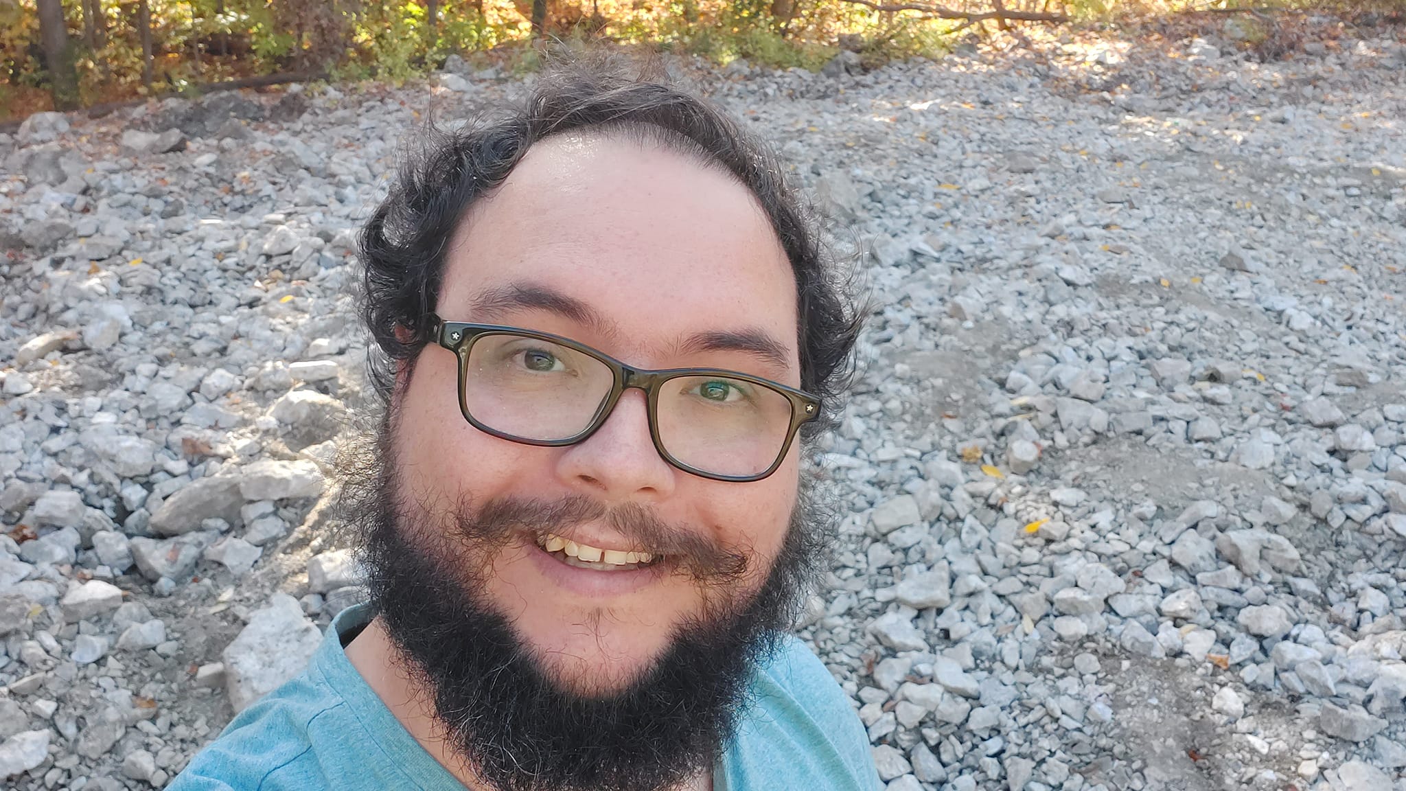 A man with short hair and bushy facial hair stands on top of a pile of off white rocks, most are pop can size but larger boulders the size of curling stones also exist